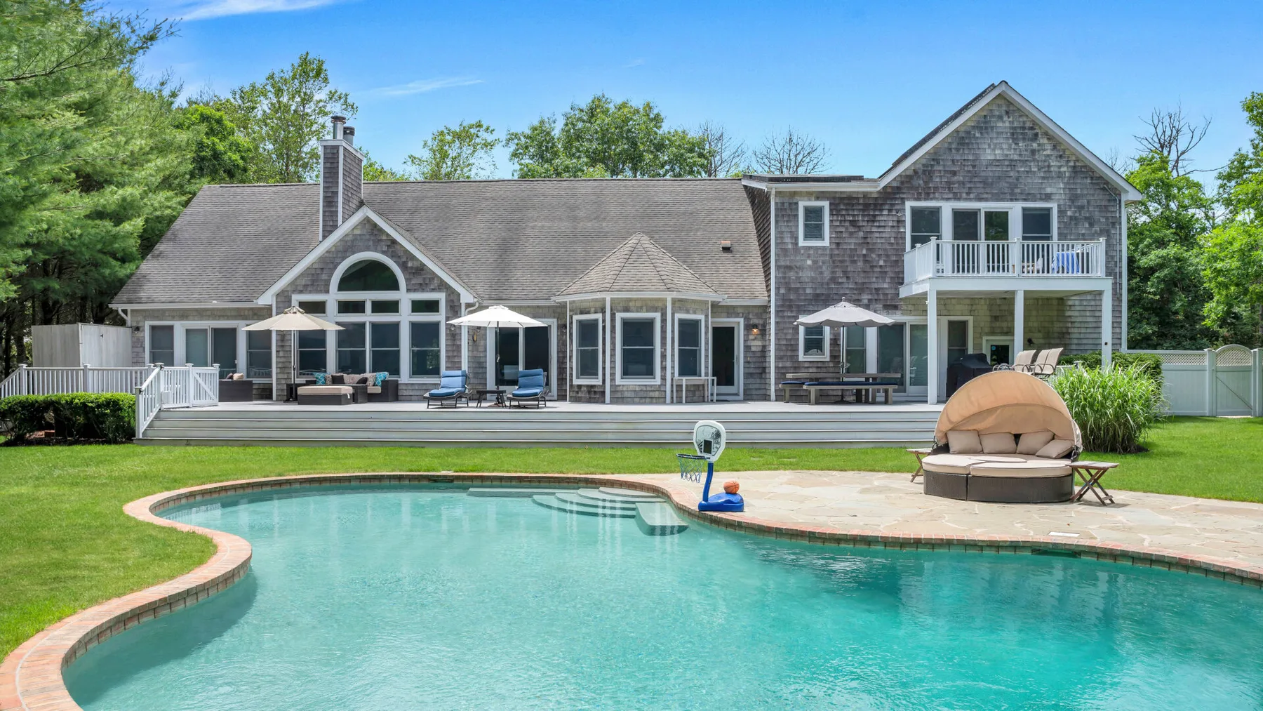 Una casa grande se alza detrás de una piscina y un patio pavimentado.