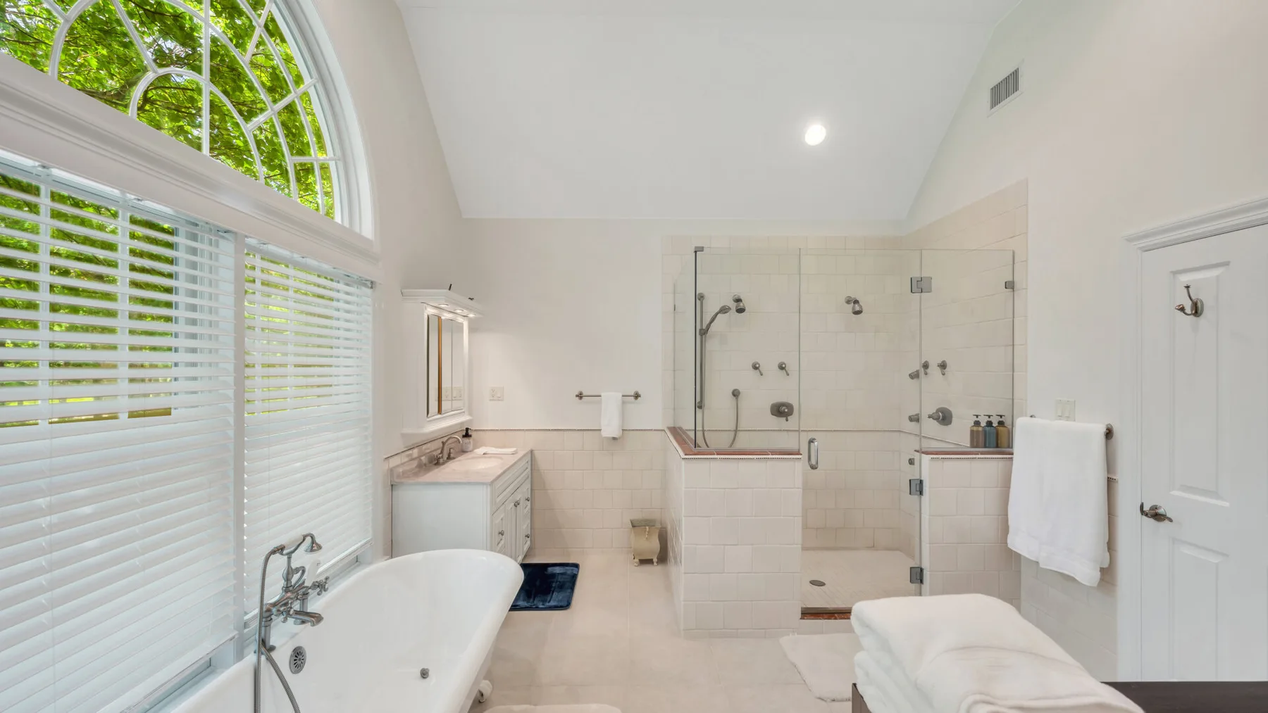 Un baño luminoso con bañera, ducha de cristal y grandes ventanas con persianas.