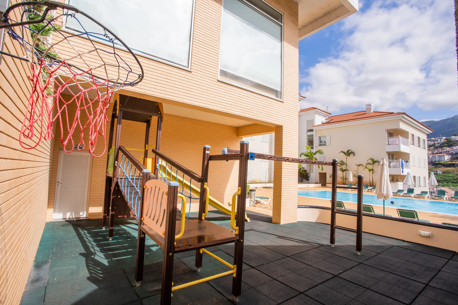 An outdoor play area with a basketball hoop overlooks a swimming pool.