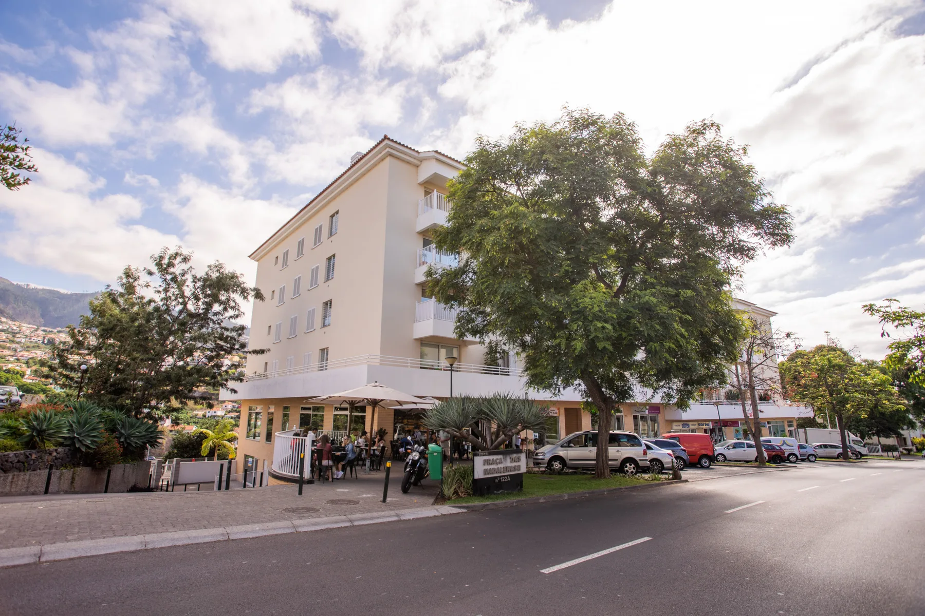 A multi-story building stands beside a tree-lined street in a sunny urban setting.