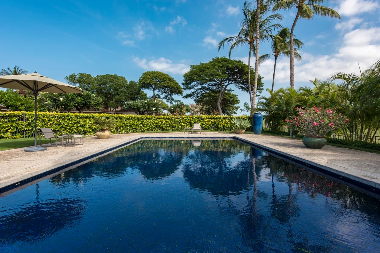 An outdoor swimming pool is surrounded by tropical plants and patio seating.