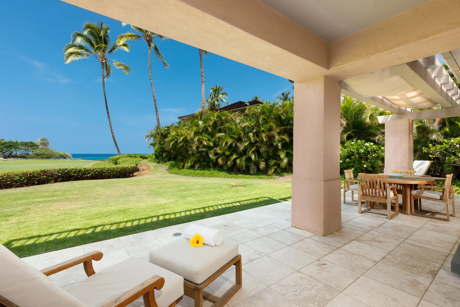 A covered patio overlooks a green lawn, tropical plants, and the ocean.