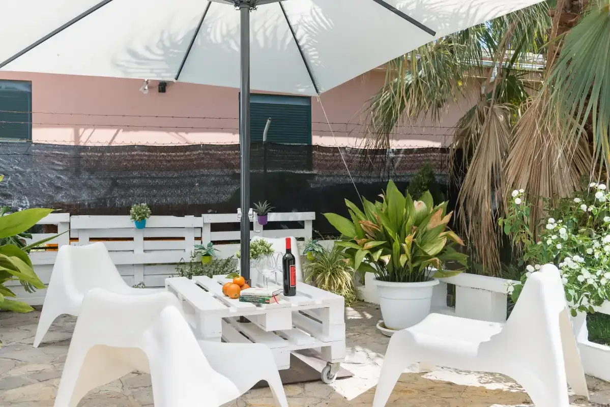 An outdoor patio has white seating, a table, and potted plants beneath an umbrella.