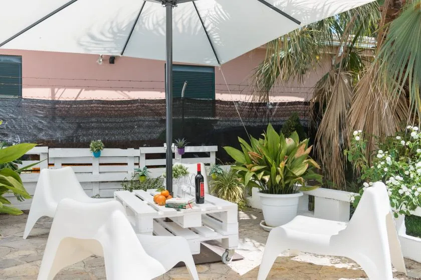 Un patio al aire libre con asientos blancos, una mesa y plantas en macetas bajo una sombrilla grande.