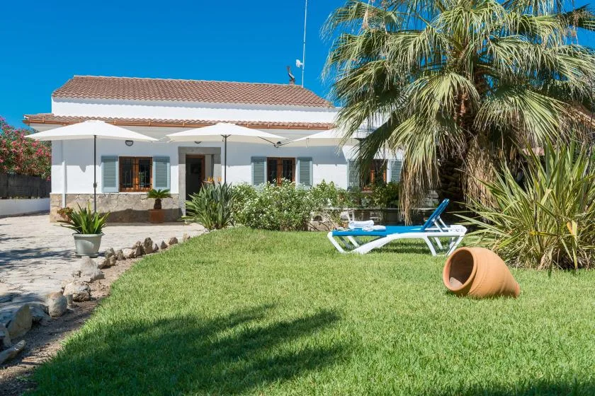 A white house overlooks a lawn with sun loungers and palm trees.
