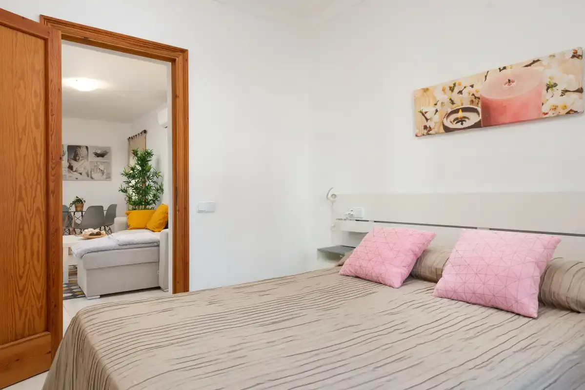 A bedroom with a bed and pink pillows, with a sitting area visible through the doorway.