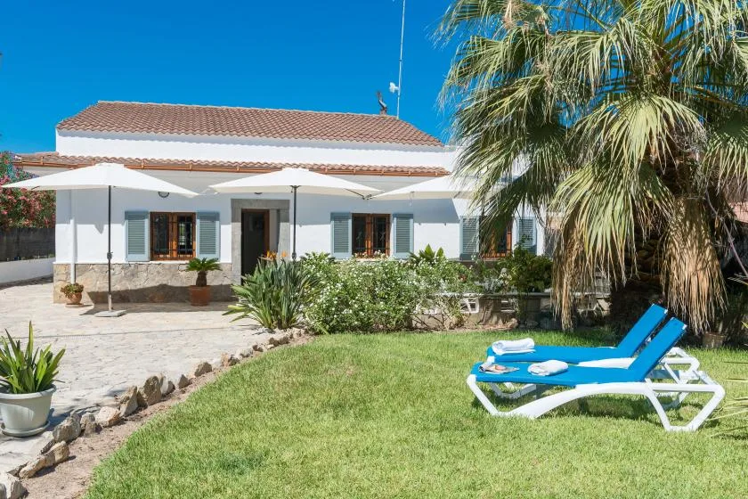 Una casa blanca da a un césped verde con tumbonas azules y una palmera.