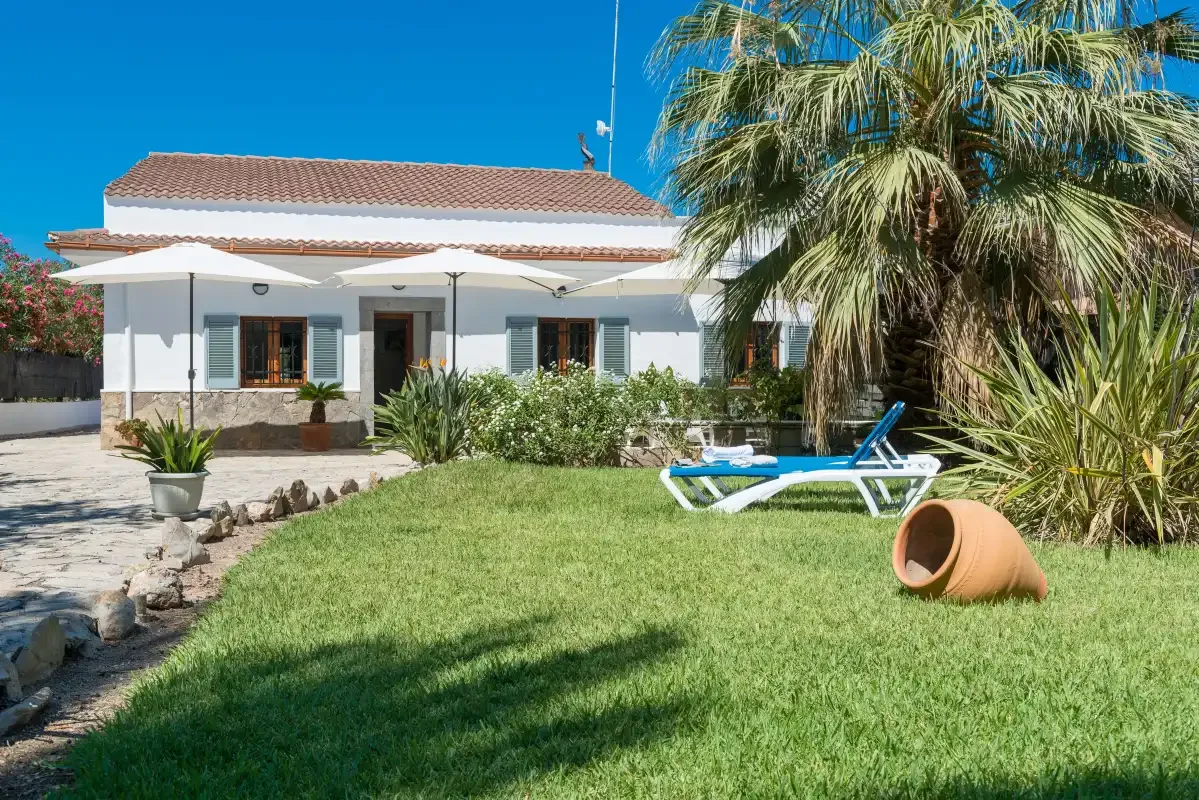 A white house faces a grassy garden with palm trees and a sun lounger.