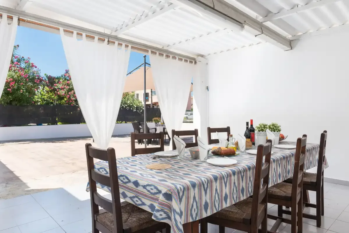 A covered outdoor dining area with a table and chairs beside an open terrace.