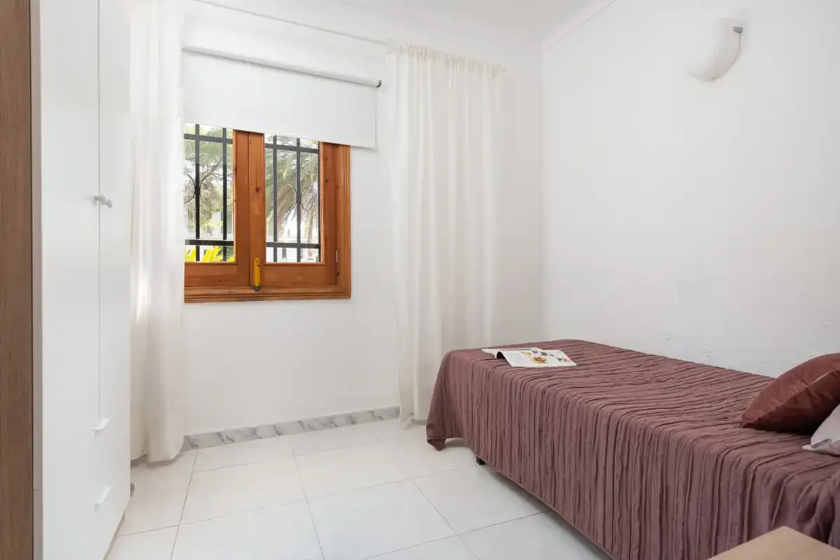 A bedroom with a bed beside a window with white curtains.