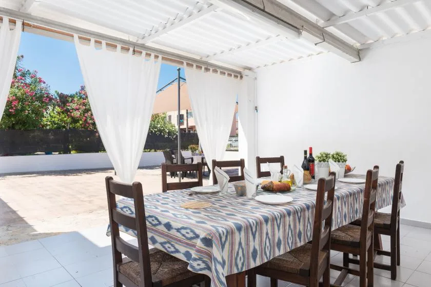 A covered outdoor dining area has a long table set with a patterned tablecloth and chairs.