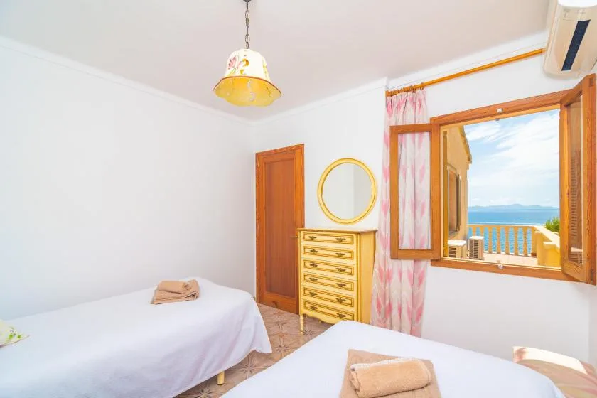 A bright bedroom with two beds, a dresser, and an open window overlooking the water.