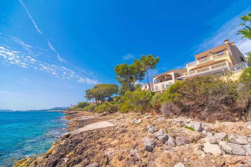 Coastal scene with rocky shoreline, clear blue water, and houses on the hillside.
