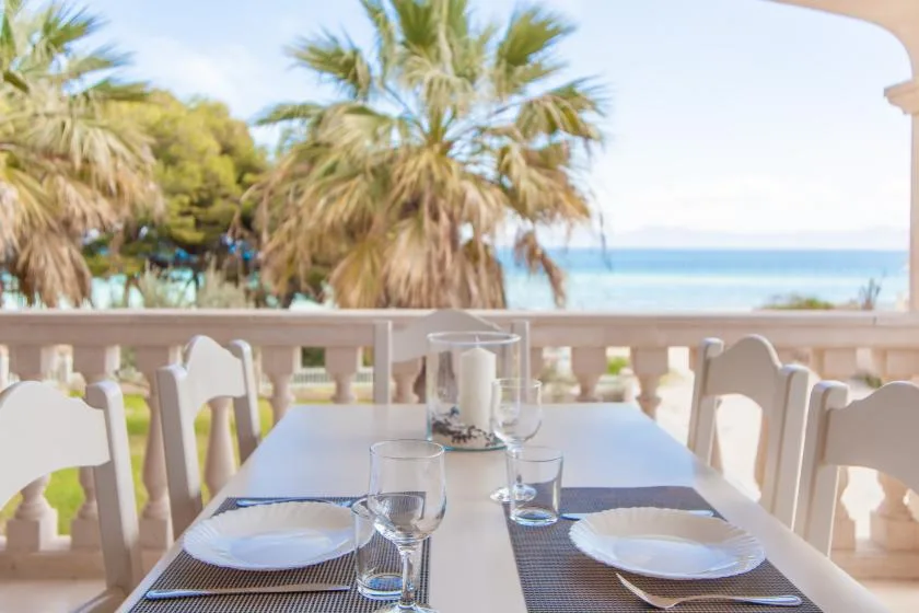 Una mesa junto al mar está preparada con platos y vasos bajo una terraza sombreada.