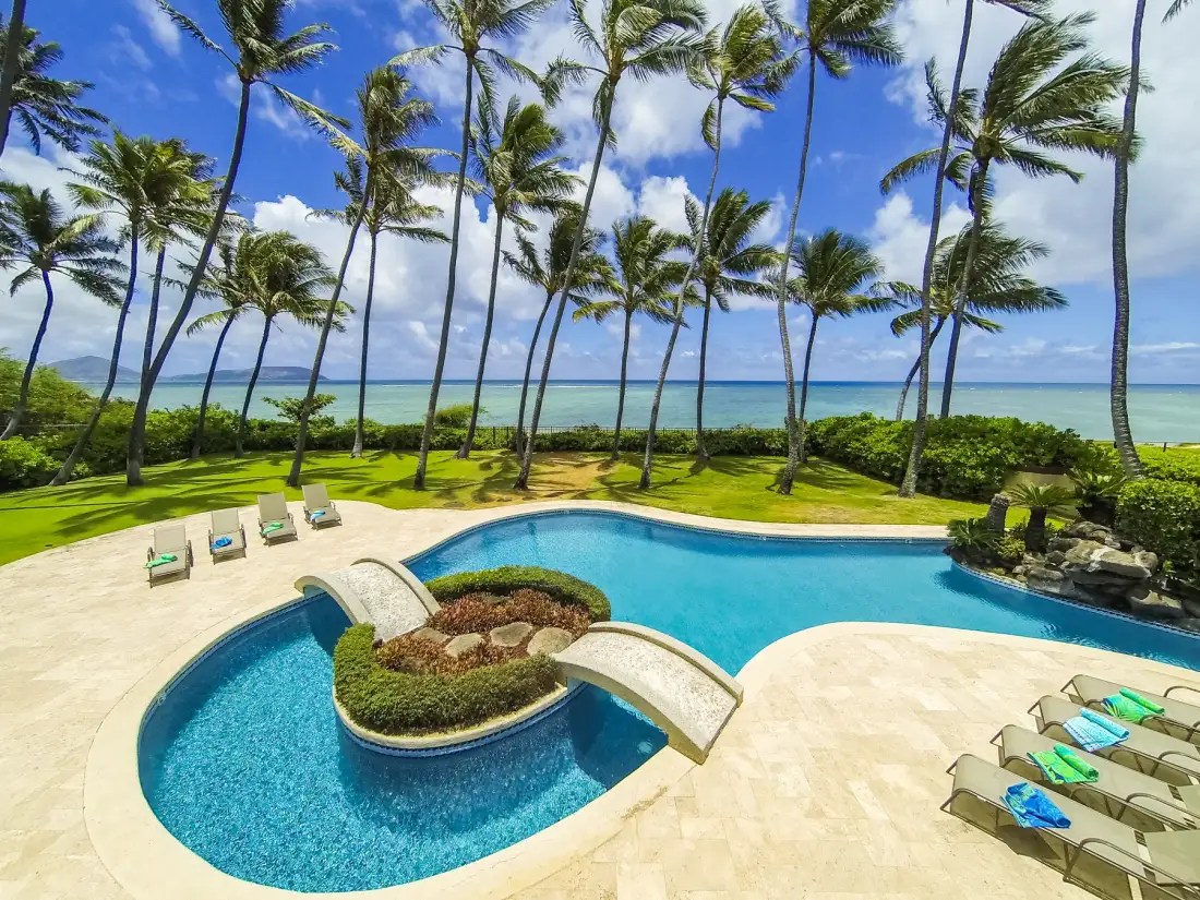 Una piscina tropical rodea una isla ajardinada, con palmeras y el océano al fondo.