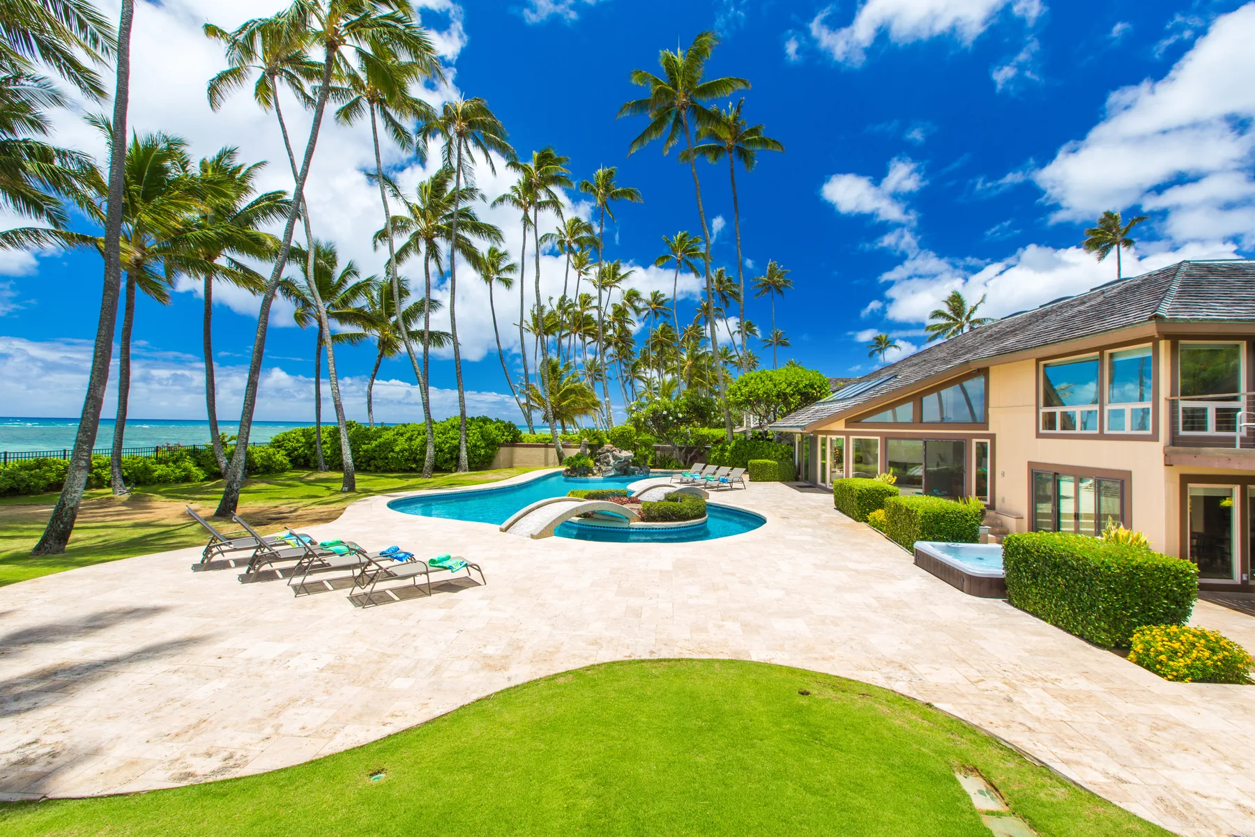 Un patio tropical con piscina junto a una casa, palmeras y vistas al océano.