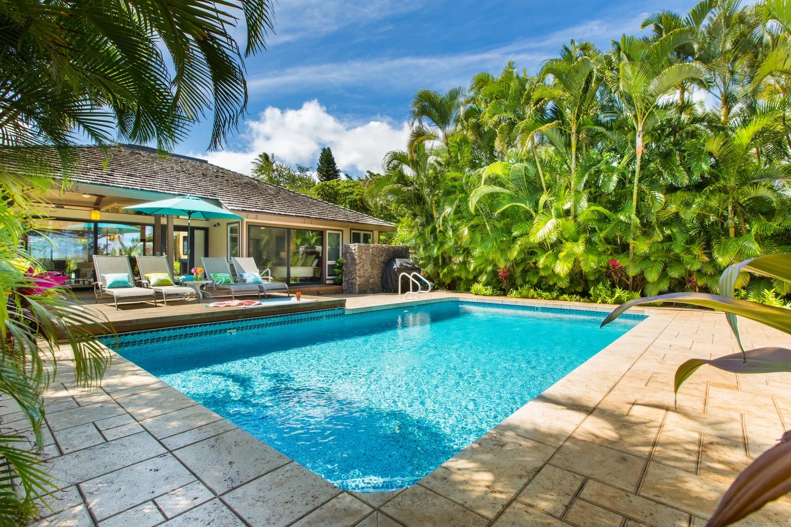 Una piscina junto a un patio de baldosas y un edificio bajo, rodeados de plantas tropicales.