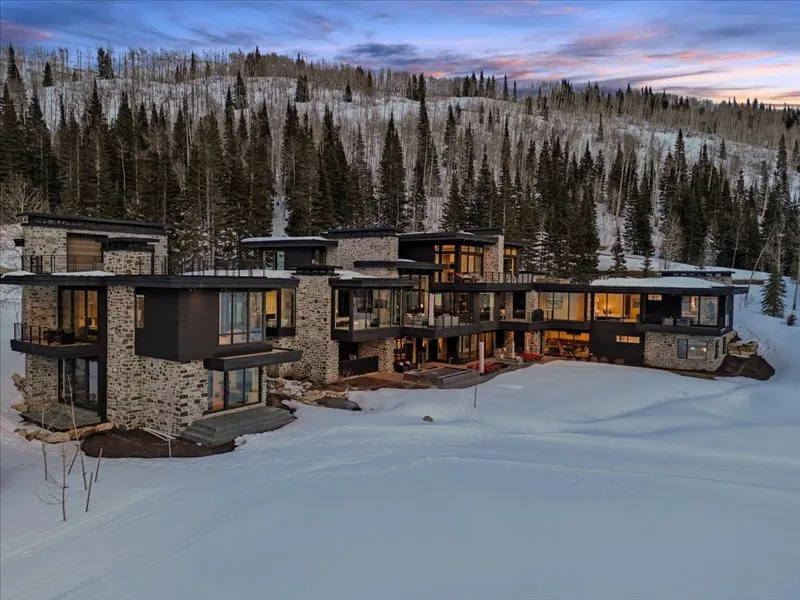 A modern mountain lodge sits below a snow-covered forest at dusk.