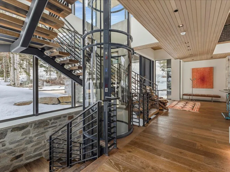 A modern interior with a central glass elevator, staircase, and wood flooring.