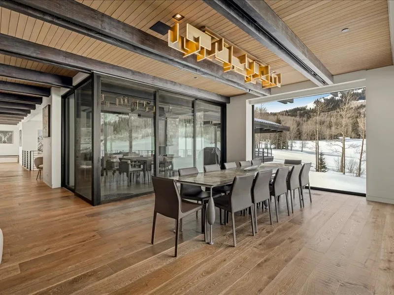 A dining area with a long table and seating beside large glass windows.