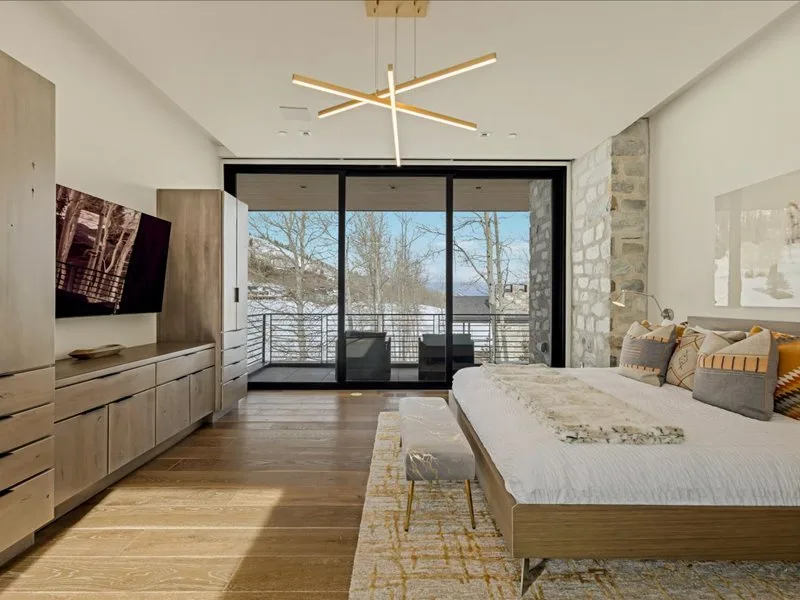 A bright bedroom with a bed, wooden furnishings, and large glass doors opening to a balcony.