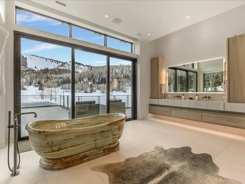 A modern bathroom has a freestanding bathtub beside broad windows overlooking a snowy landscape.