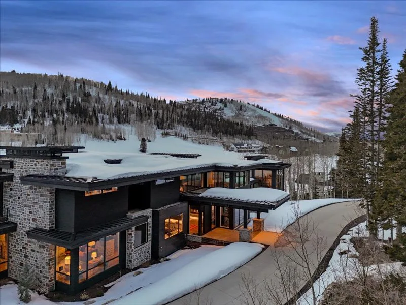 A modern mountain home sits beside a snow-covered hillside at dusk.