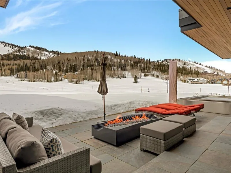A covered outdoor seating area overlooks a snowy mountain landscape, with a fire pit in front.