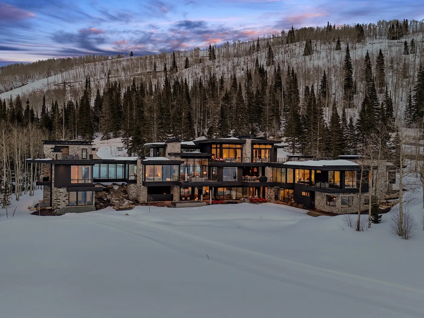 A modern building sits in a snowy landscape beneath a forested hillside at dusk.