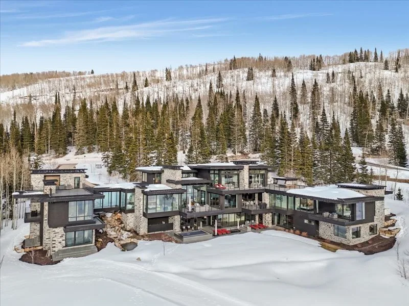 Modern buildings sit in a snow-covered landscape beneath a forested hillside.