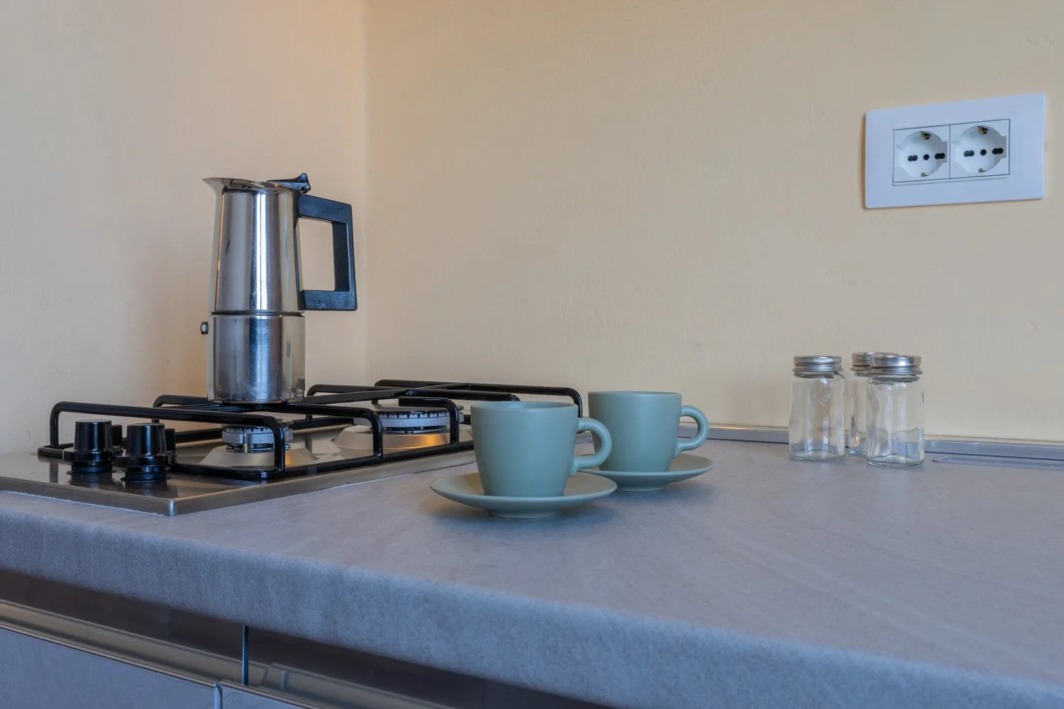 Una encimera de cocina con una cafetera de estufa, dos tazas y frascos de vidrio.