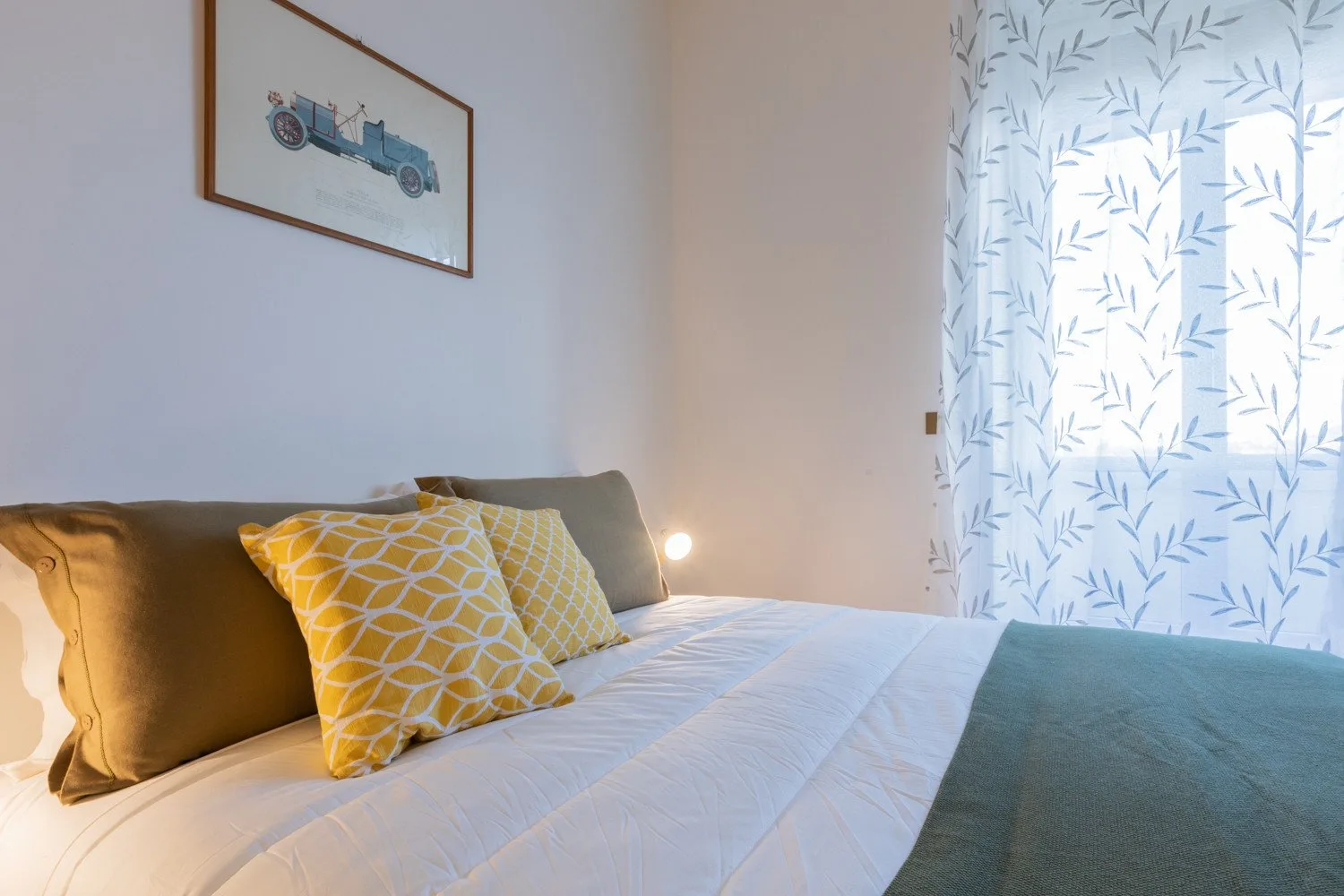 Un dormitorio con una cama, cojines estampados y una ventana con cortinas claras.