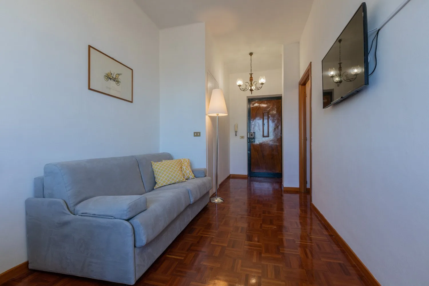 Un pasillo estrecho con zona de estar tiene asientos grises, una lámpara de pie y una puerta de madera.