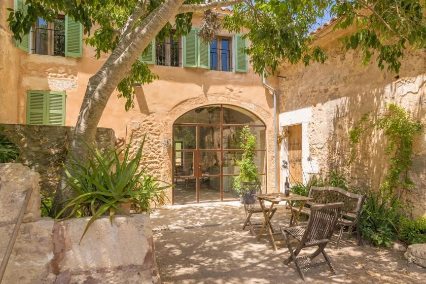 A sunlit courtyard with a glass entrance, leafy plants, and outdoor seating.