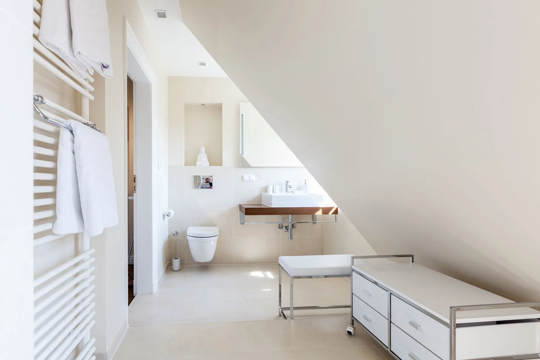 Baño luminoso con lavabo, inodoro y radiador toallero bajo un techo inclinado.