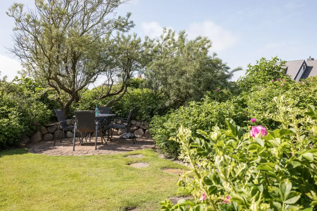 Zona de estar en un jardín, con una mesa bajo árboles frondosos y arbustos en flor.