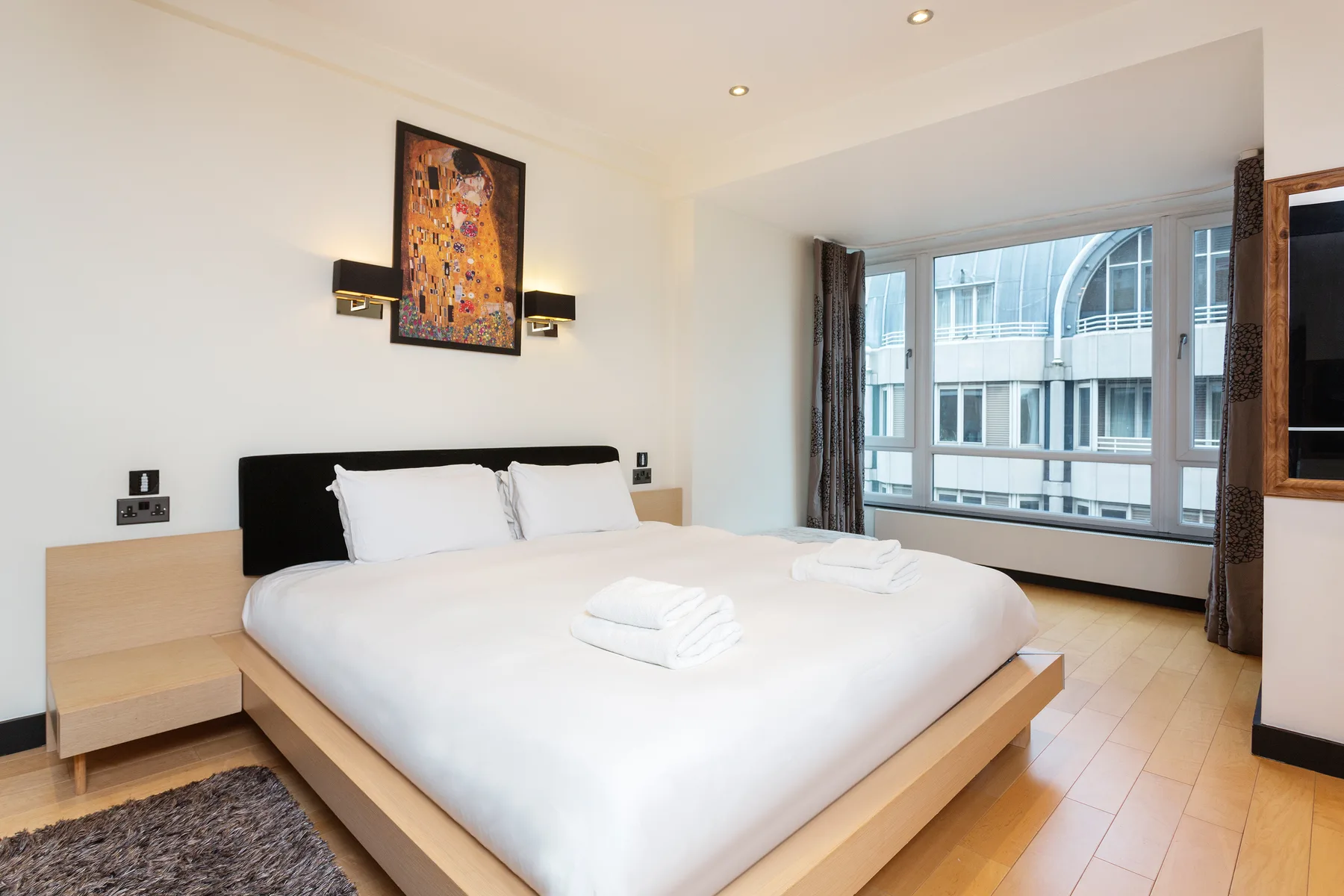 A bright bedroom with a large bed and wide windows overlooking nearby buildings.