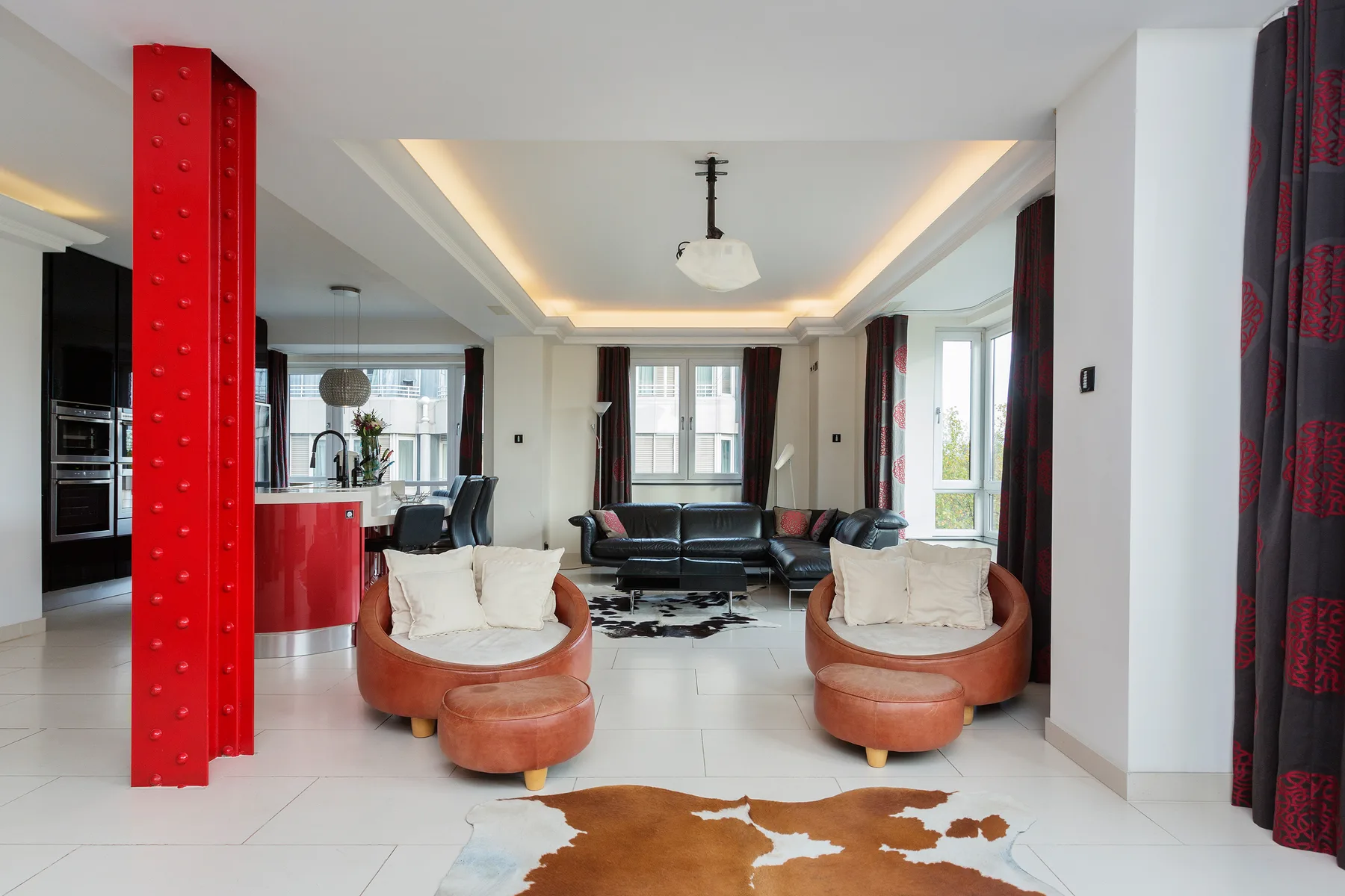 A bright open-plan living room with seating, a red kitchen counter, and large windows.