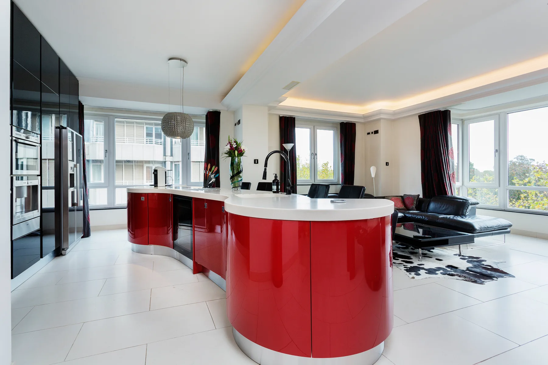 A bright open-plan kitchen and living area with a curved red island and large windows.