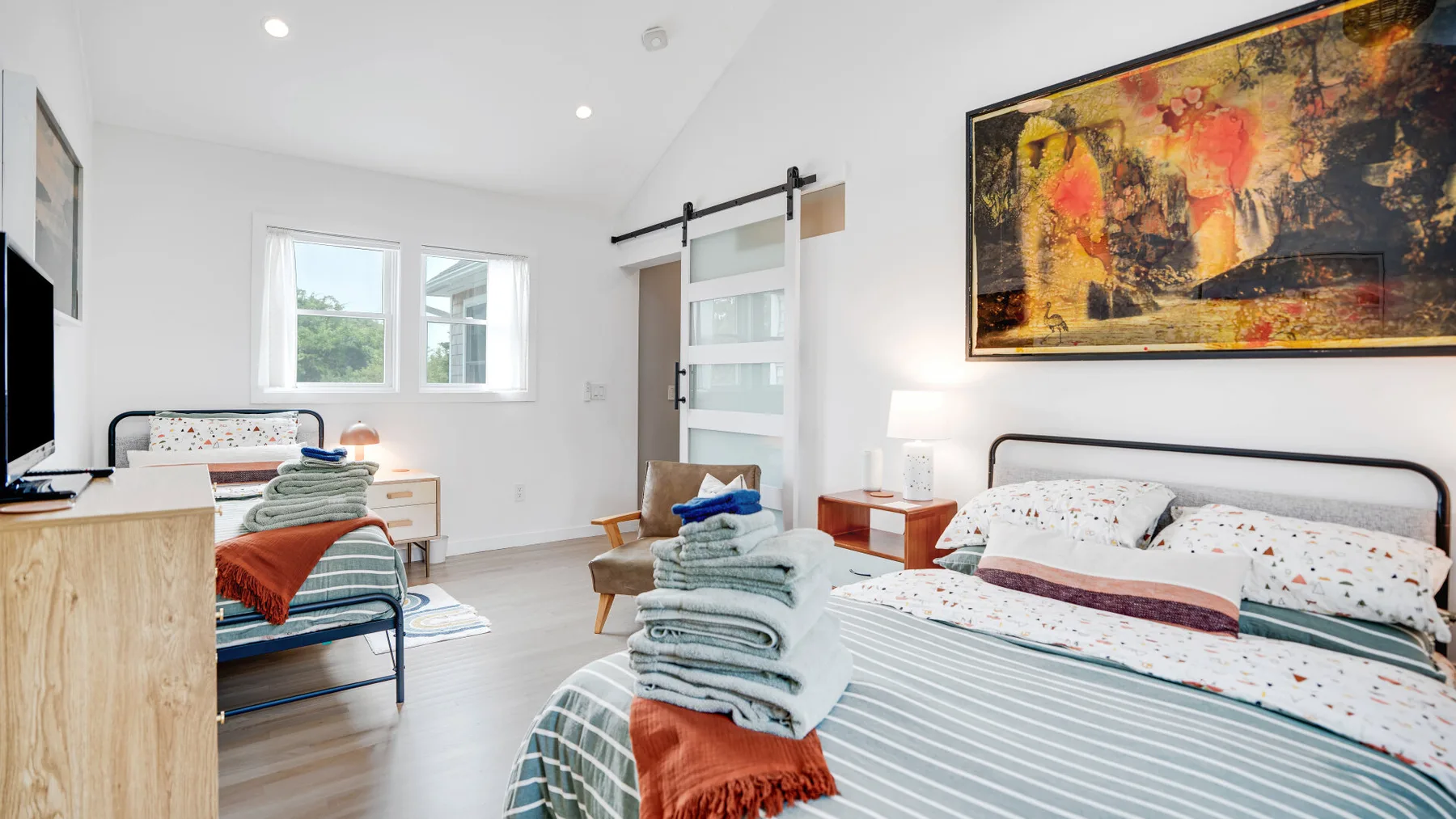 Un dormitorio luminoso con una cama, una pequeña zona de estar y arte colorido en la pared.