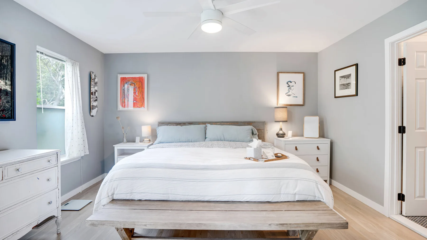 Un dormitorio luminoso con una cama, mesitas de noche y cuadros enmarcados.
