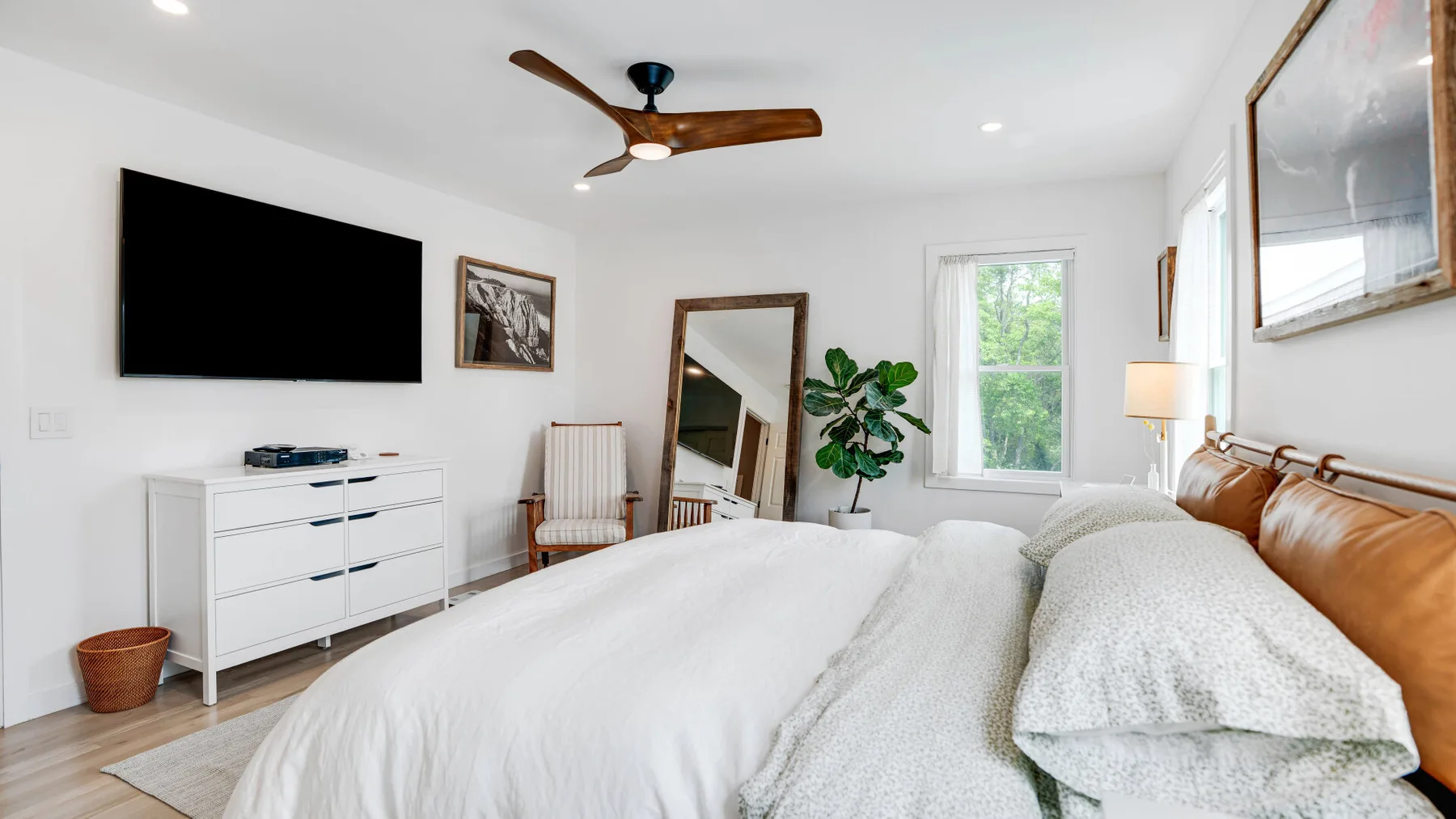 Un dormitorio luminoso con ropa de cama blanca, una cómoda y una ventana con vistas a la vegetación.