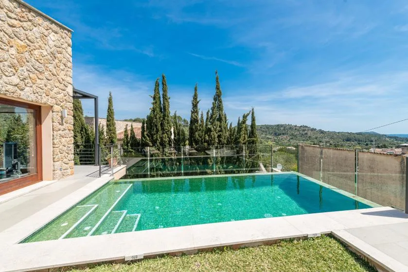 Outdoor swimming pool beside a stone building, with trees and hills in the background.