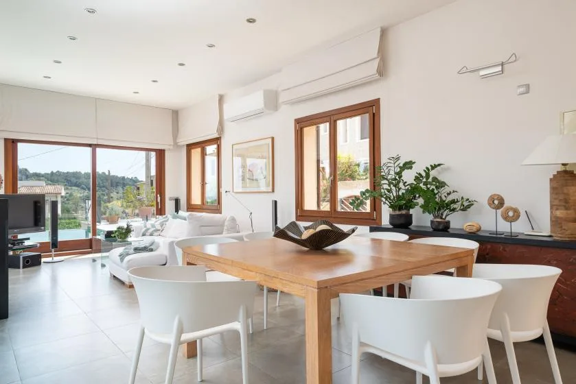 A bright dining and living area features a wooden table and large windows.