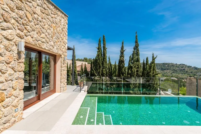 A swimming pool beside a stone house, with trees and hills in the background.