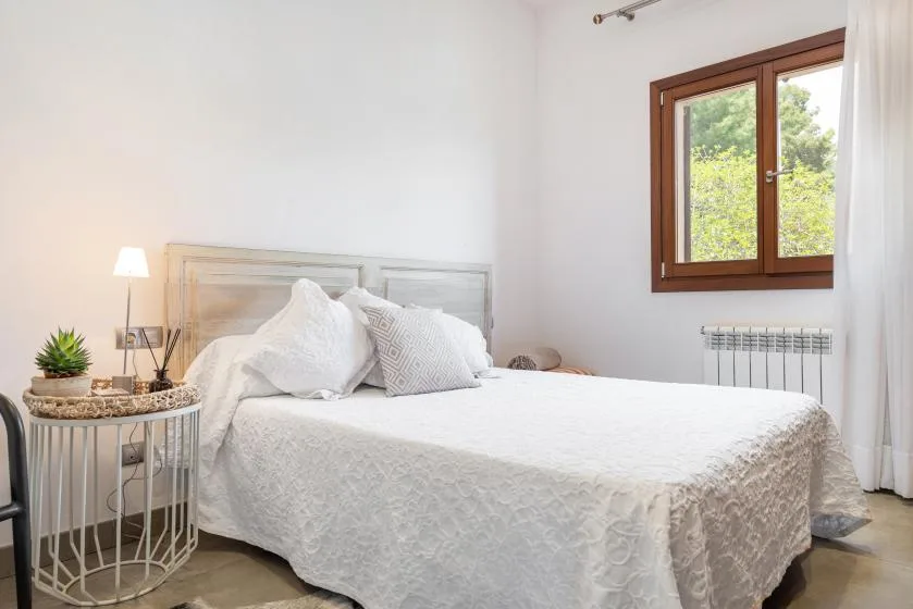 A bright bedroom with a bed dressed in white bedding beside a window.