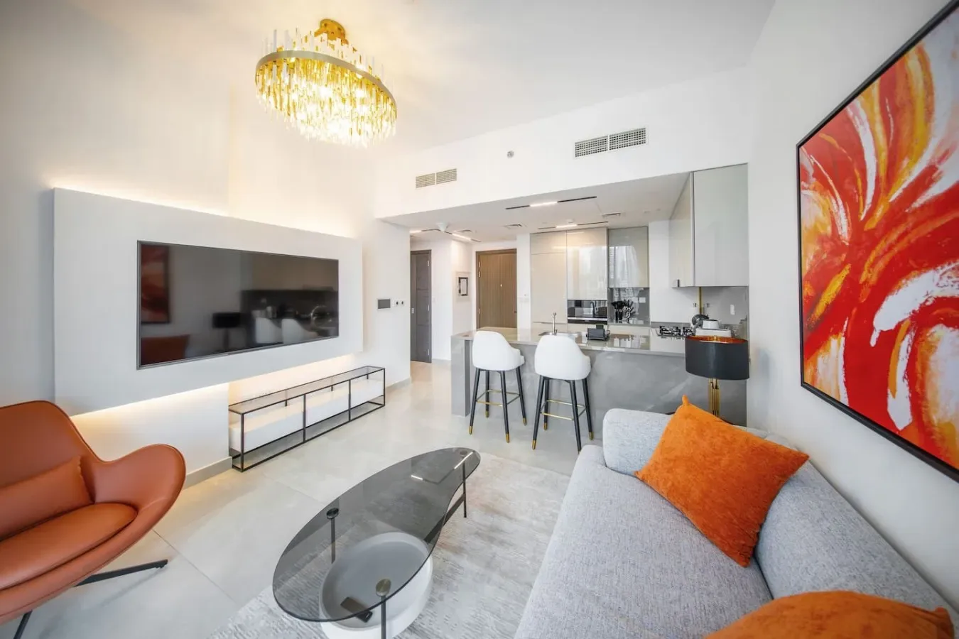 A bright living room opens onto a kitchen, with a wall-mounted television and colorful artwork.