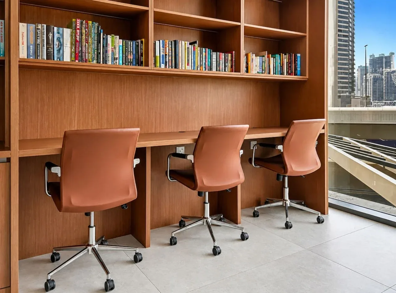 A study area with a long wooden desk, bookshelves, and seating beside large windows.