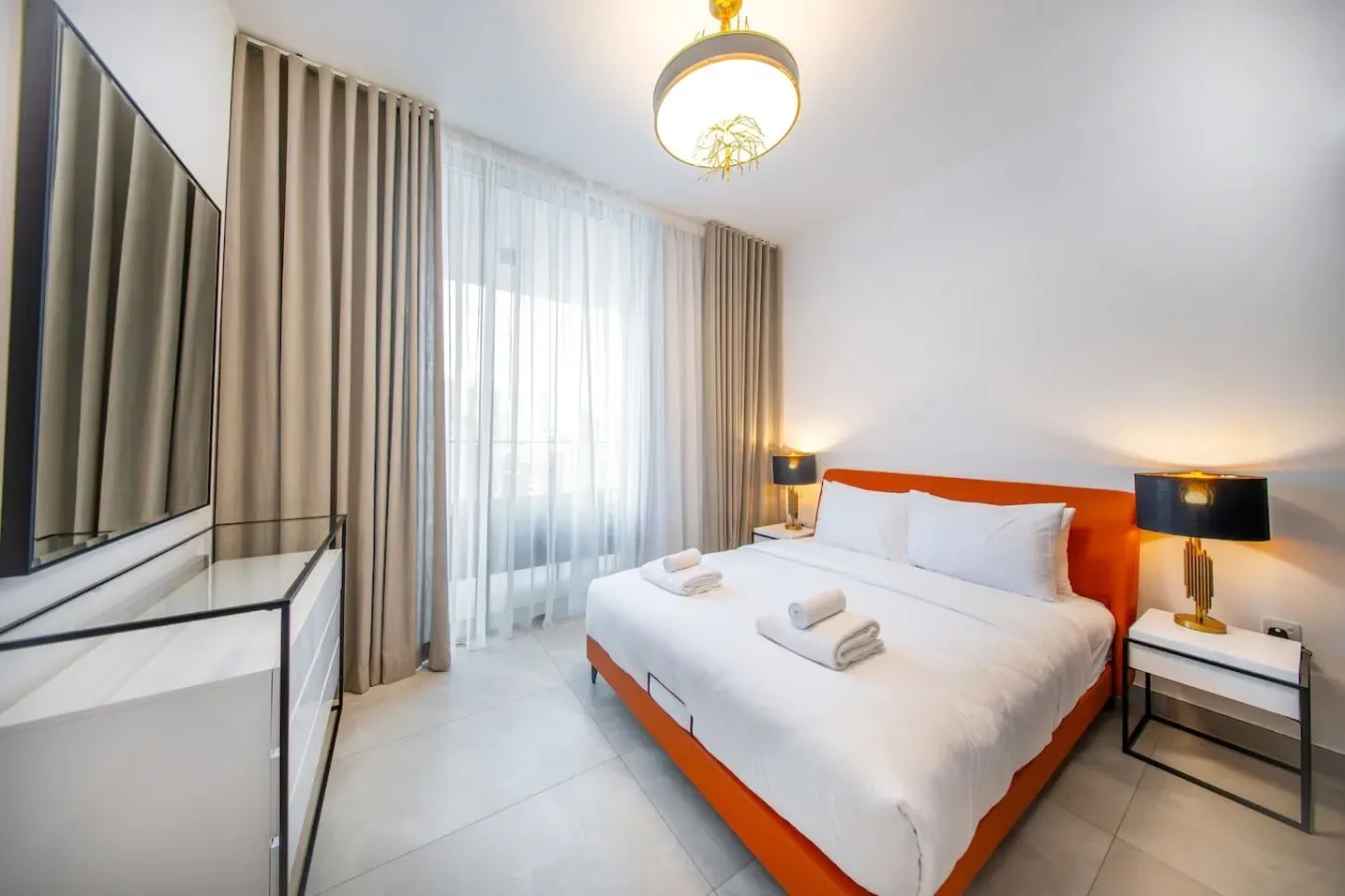A bright bedroom with a bed, curtained window, and television.