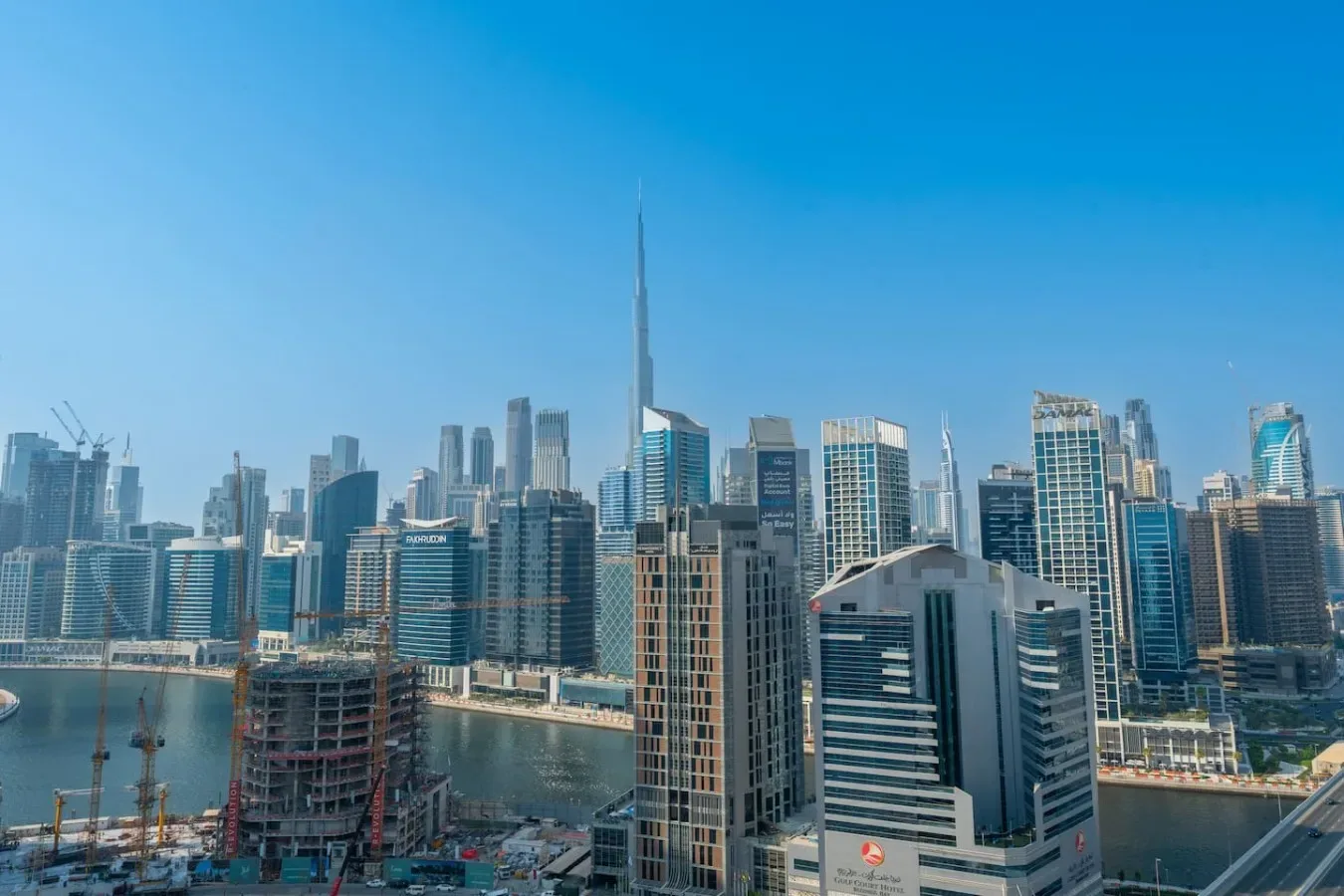 A city skyline of tall buildings rises beside a waterfront under a clear blue sky.